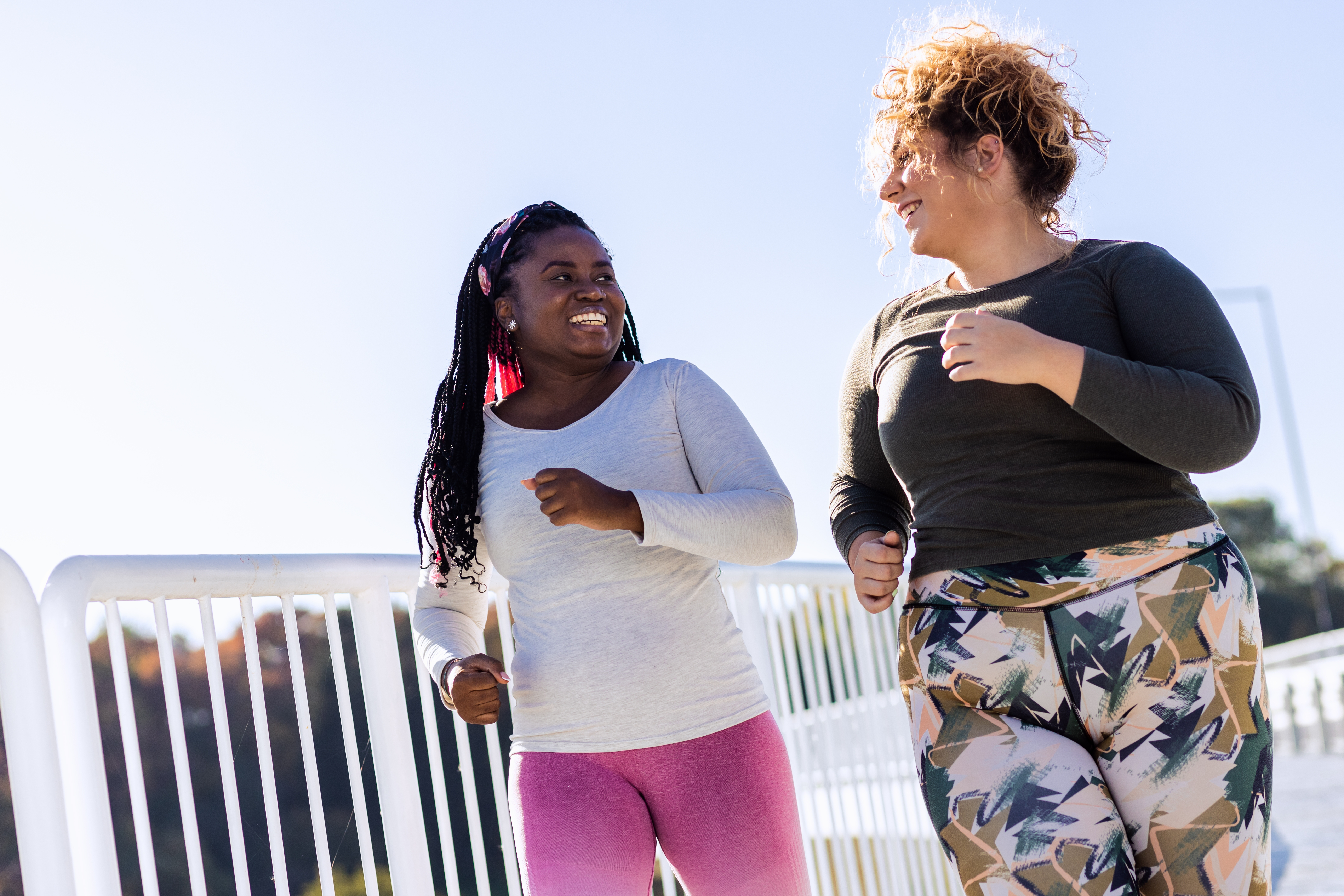 Two smiling women jogging outdoors on a sunny day, wearing long-sleeve tops
and colorful leggings